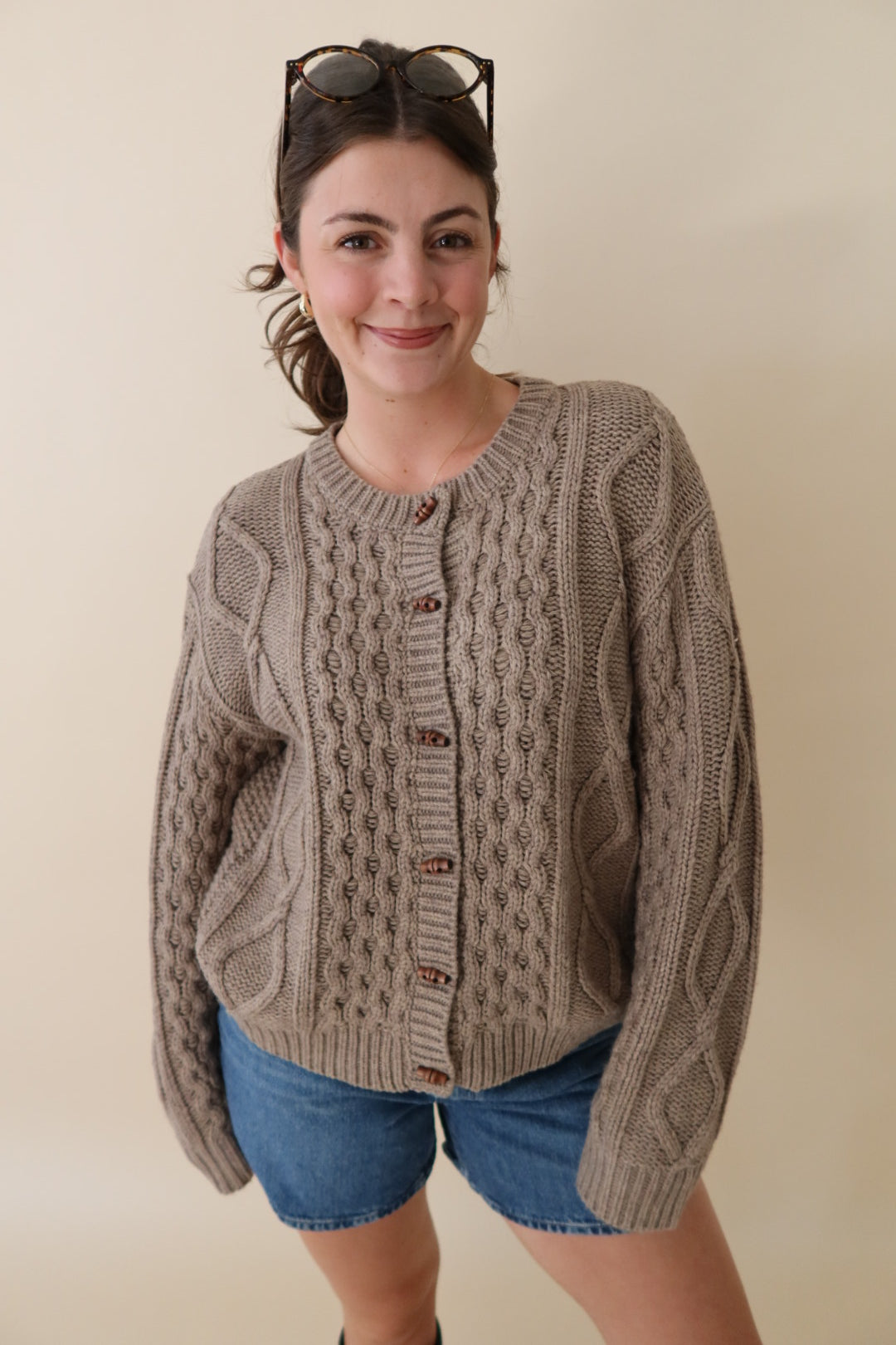 A woman wearing a chunky cable-knit button-front cardigan in taupe-beige with brown buttons, paired with denim shorts and sunglasses on her head, against a plain beige background.