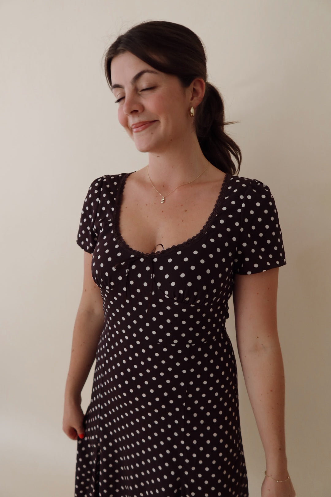 A woman wearing a black short-sleeve dress with a white polka dot pattern, standing against a plain beige wall.